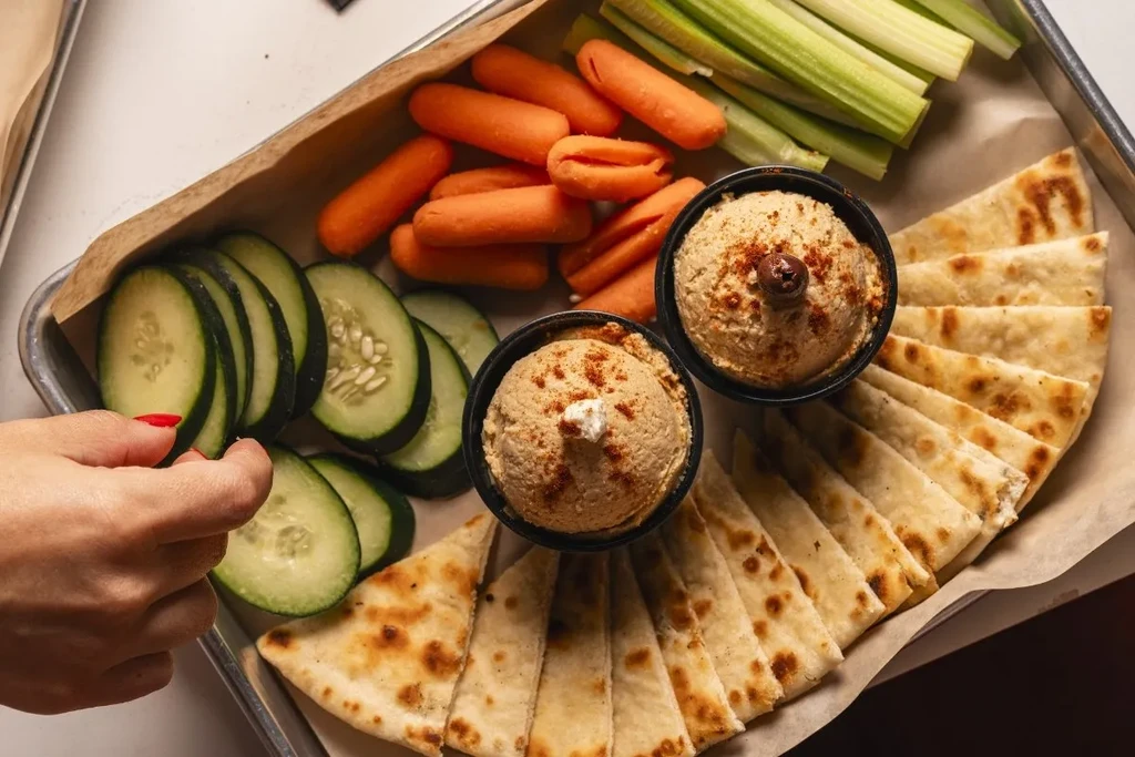 Overhead shot of two hummus bowls, crudités, pita, and a hand.