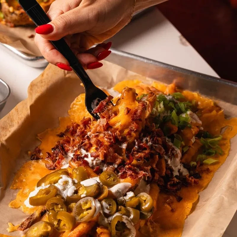 Hand scooping loaded fries or nachos with cheese, bacon, jalapenos, and sauce.