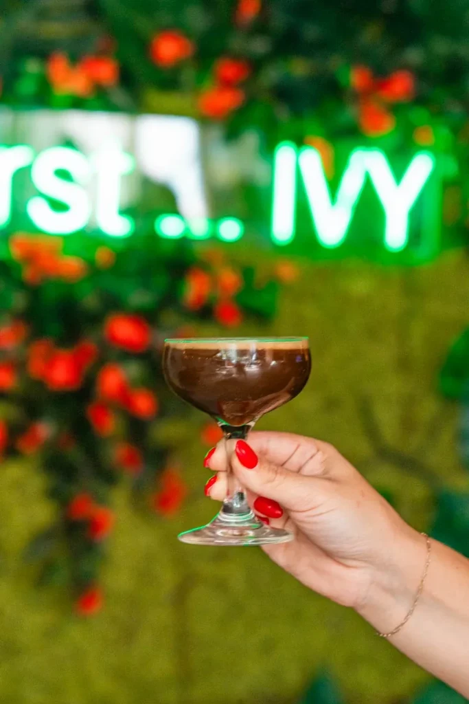 Red-nailed hand holds dark cocktail near blurred 'rst... IVY' neon sign.