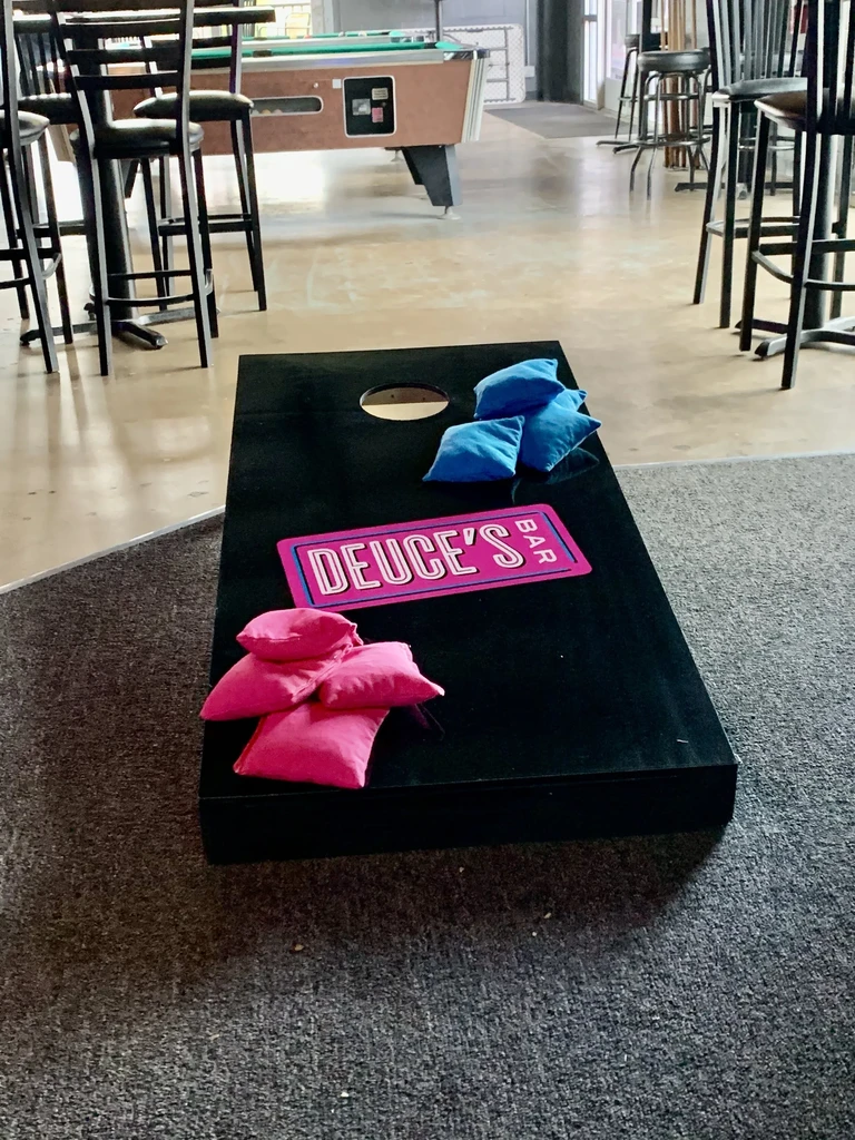 Black cornhole board with beanbags and "DEUCE'S BAR" sign in a bar.