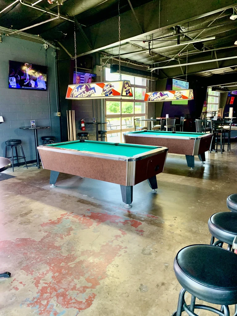 Two green-felt pool tables in an industrial bar, with Pabst lights.
