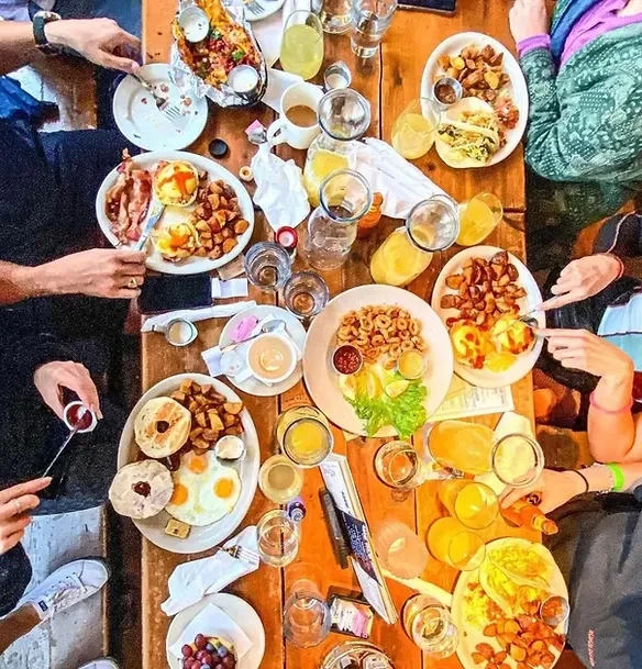 Overhead view: Many hands share a large, bustling breakfast spread.
