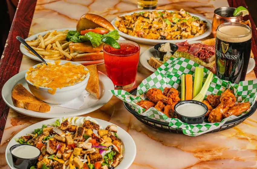 Diverse food and drinks on a marble table: burgers, wings, beer, salad.