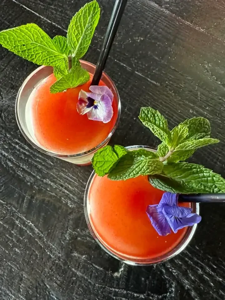 Two top-down reddish-orange cocktails with mint, purple flowers, and straws.