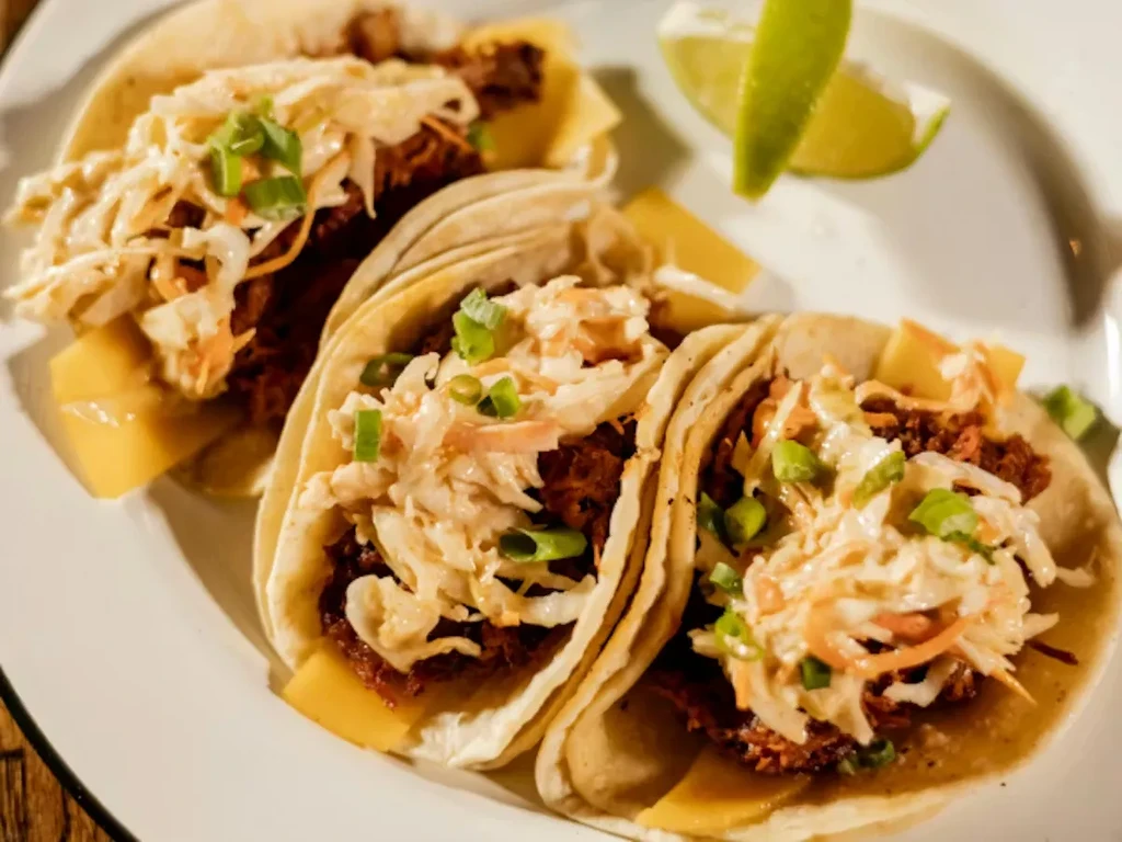 Close-up of three dark meat tacos topped with slaw, fruit, and lime.