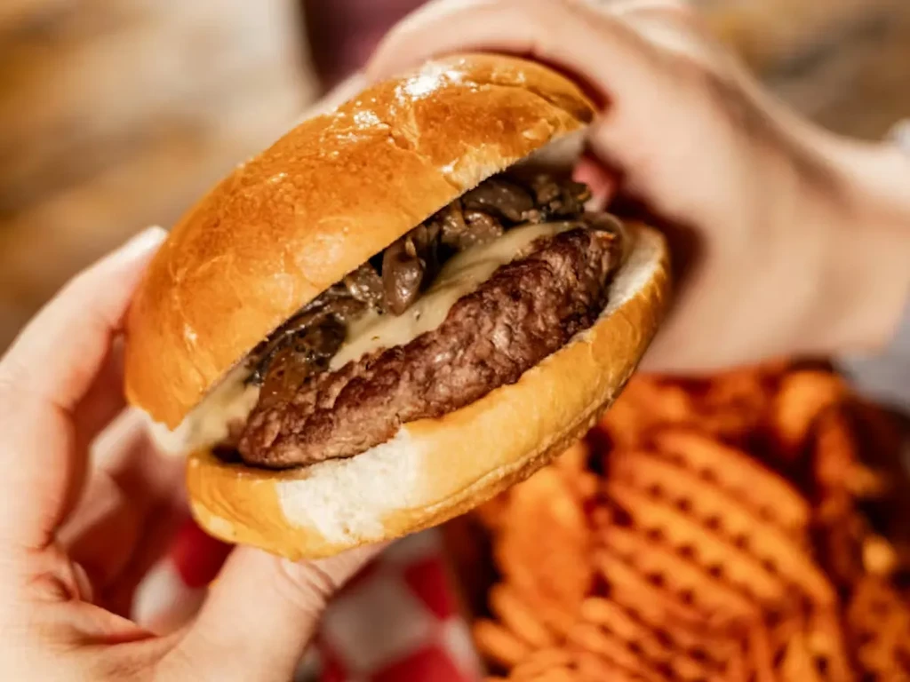 Hands holding a gourmet mushroom and cheese burger near waffle fries.