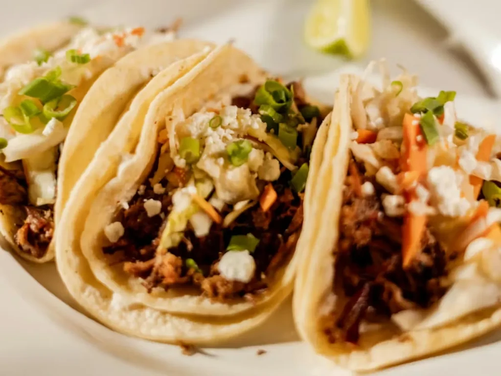 Three tacos with shredded meat, cheese, slaw, scallions, lime.
