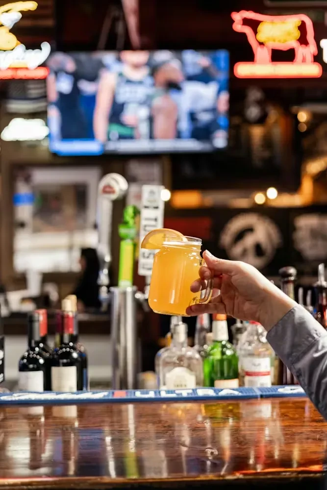 Hand with citrus drink in mason jar, blurred bar background.