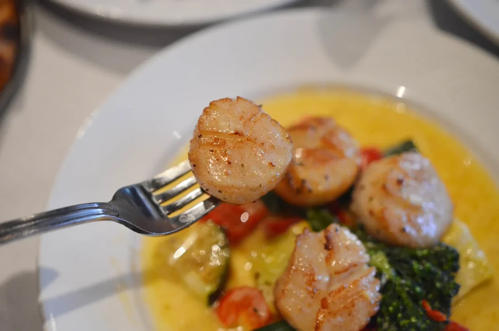 Seared scallop on a fork, with scallops, yellow sauce, and vegetables.