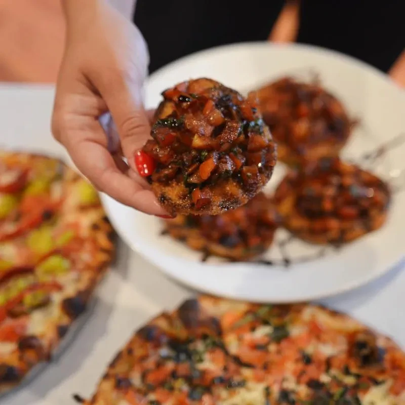 Hand holding tomato bruschetta; blurred wood-fired pizzas and appetizers.