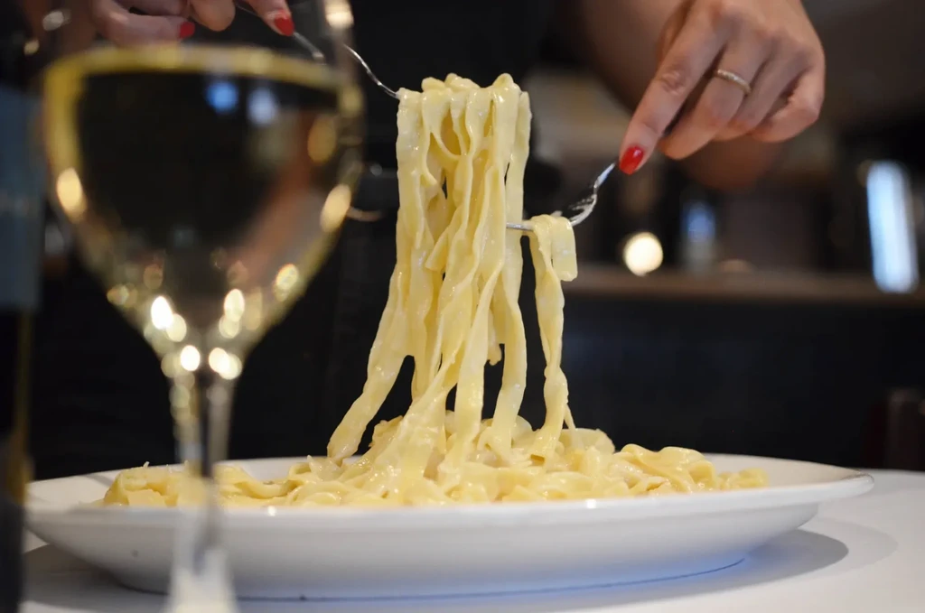 Hands twirl creamy pasta onto forks; blurred wine glass foreground.