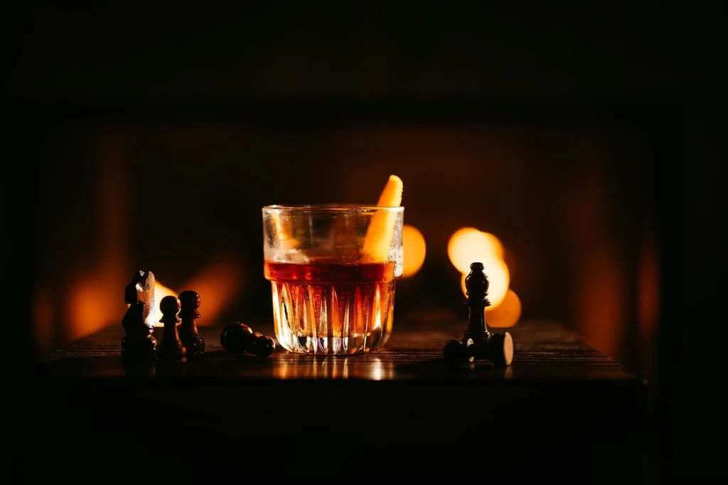 Amber cocktail with citrus peel, surrounded by dark chess pieces.