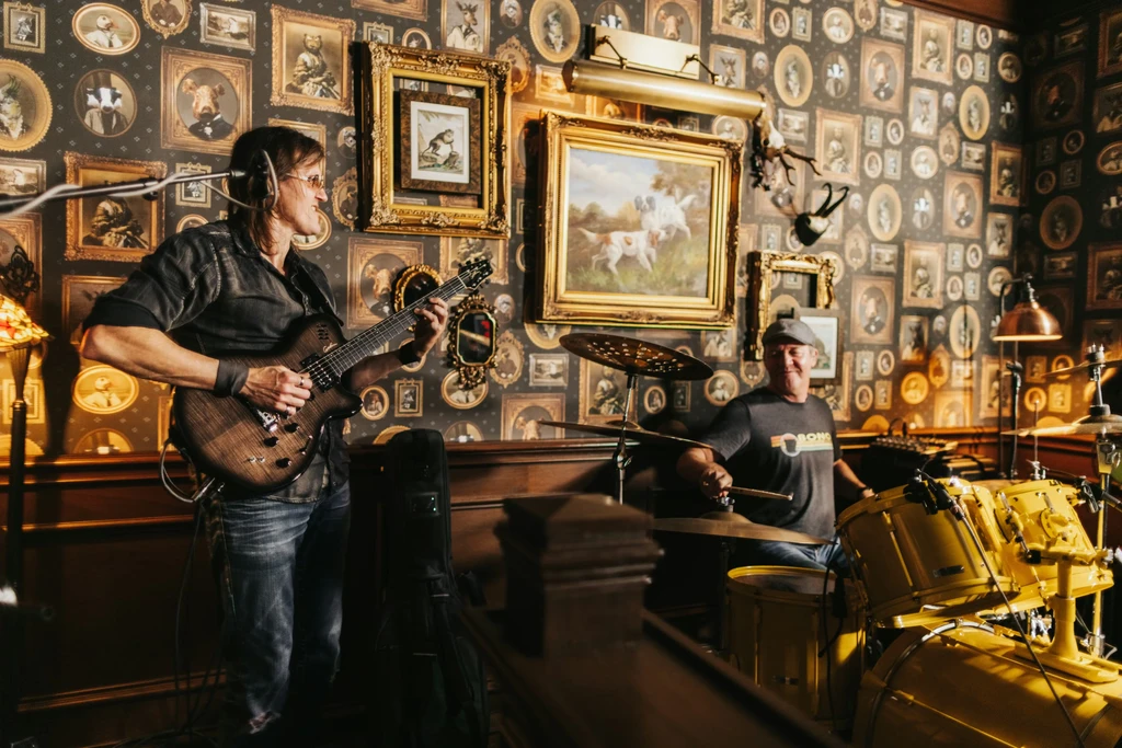 Two men play electric guitar and drums in an eclectic room.