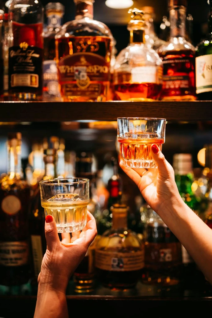 Hands toasting amber drinks with ice, liquor bottles blurred behind.
