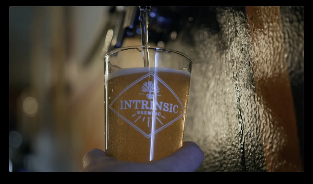 Close-up: Golden beer fills "INTRINSIC BREWING" glass from a tap.