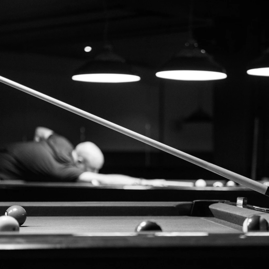 Black and white: foreground pool cue, balls, person in dim hall.