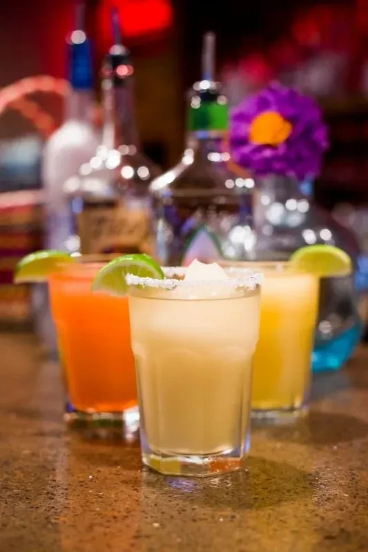 Three colorful cocktails with lime wedges on a dark, blurred bar.