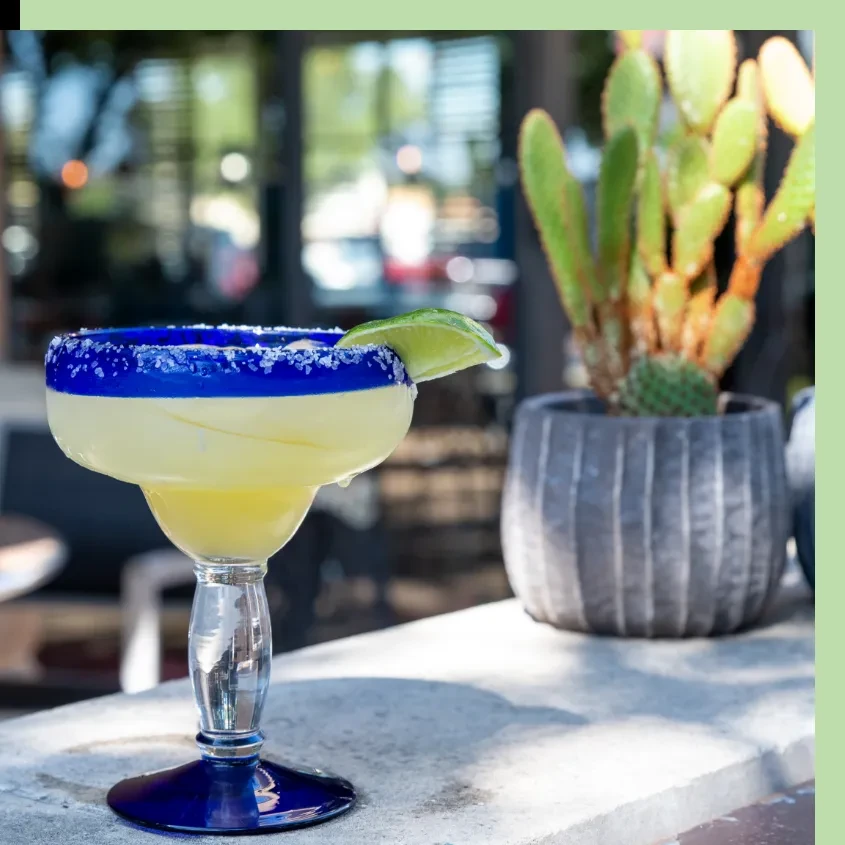 Bright yellow margarita in blue-rimmed glass with lime wedge, cactus background.