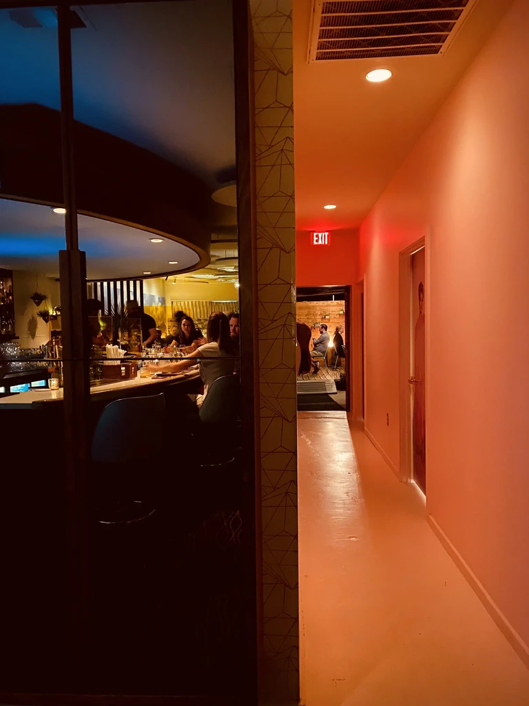Dark bar with patrons, bright pink hallway with EXIT sign.