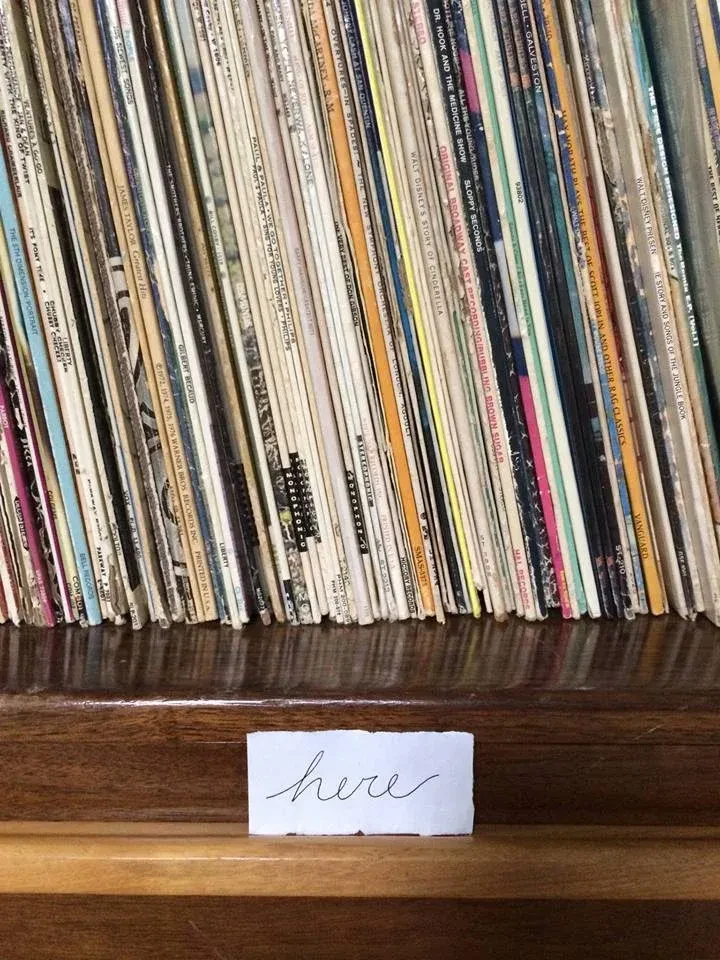 Spines of stacked vintage vinyl records and a 'here' note on wood.
