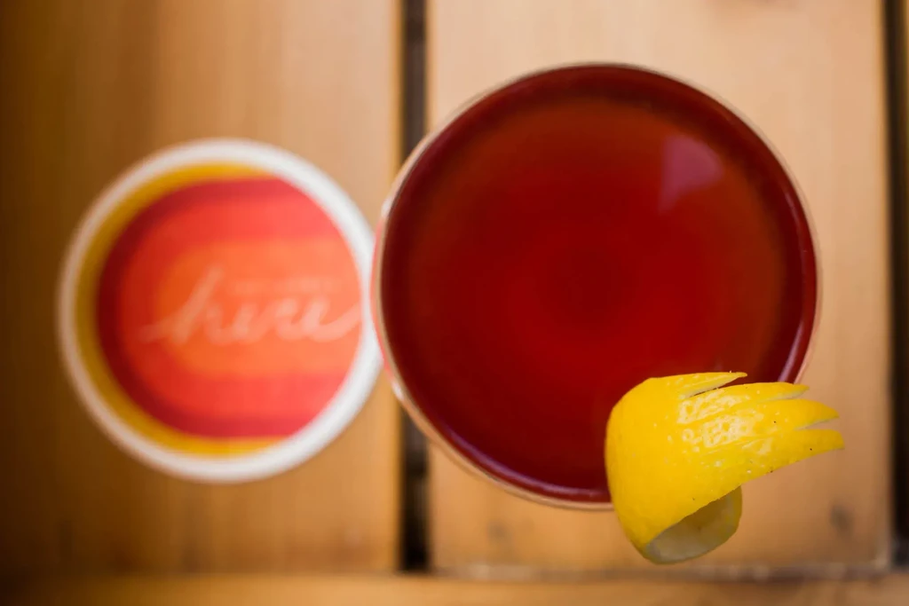 Dark red cocktail with yellow spiraled lemon peel on blurred coaster.