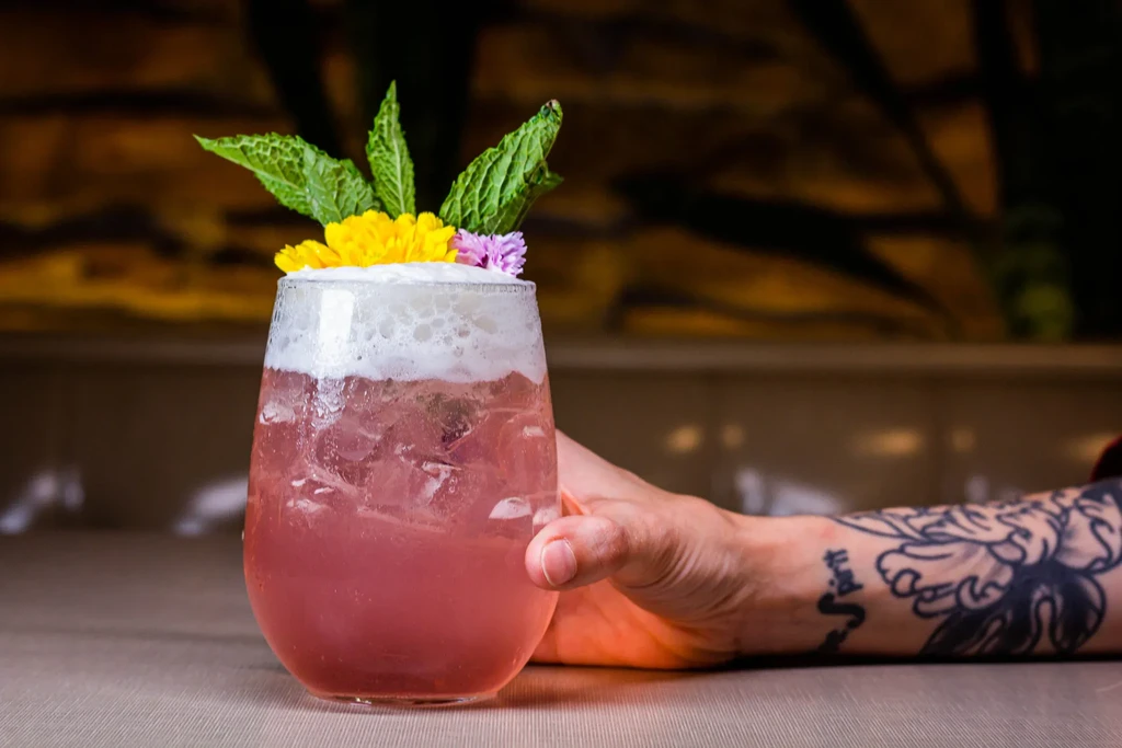 Pink cocktail with foam, mint, and flowers, held by a tattooed hand.