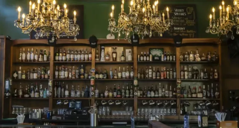A bar's liquor shelves and counter beneath ornate chandeliers on a green wall.