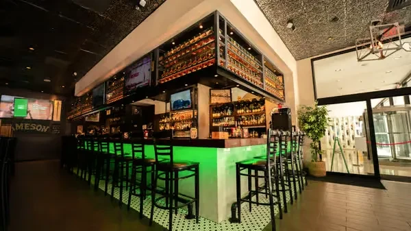 Empty modern bar with illuminated green counter, liquor shelves, and TVs.