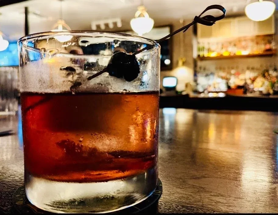 Amber cocktail with a cherry on a pick, on a blurred bar.