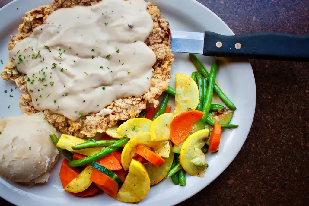 Oval plate with country fried steak, gravy, mashed potatoes, and vegetables.