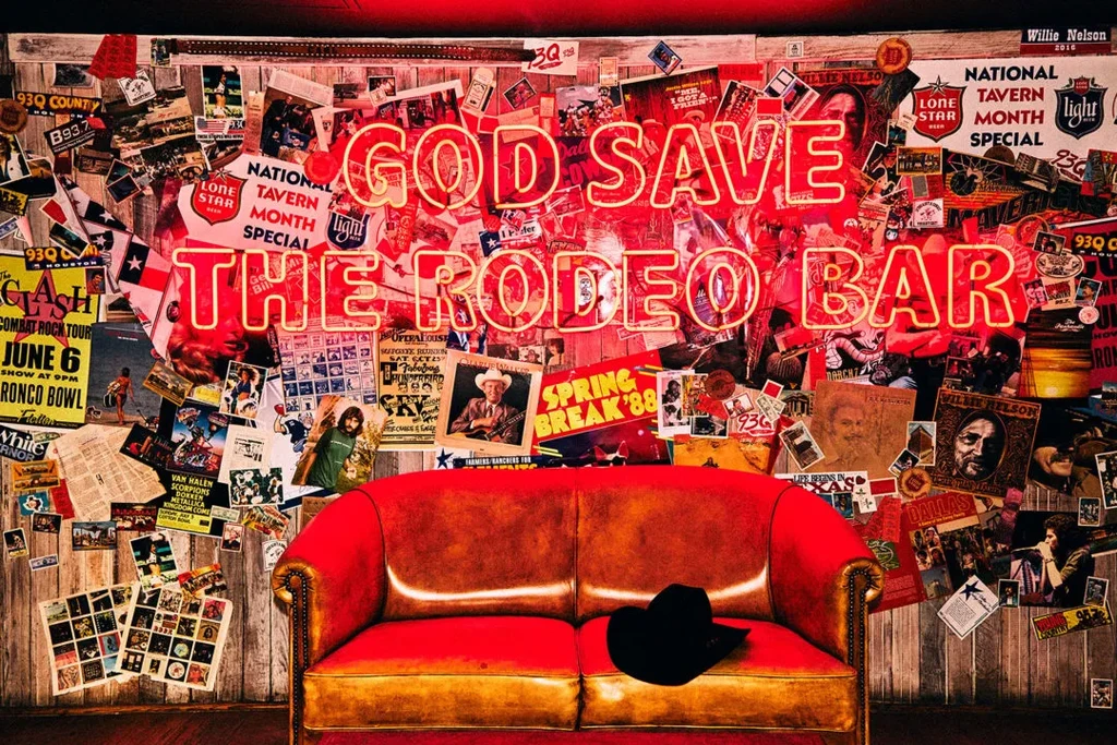 Vintage poster wall with "GOD SAVE THE RODEO BAR" neon sign and red couch.