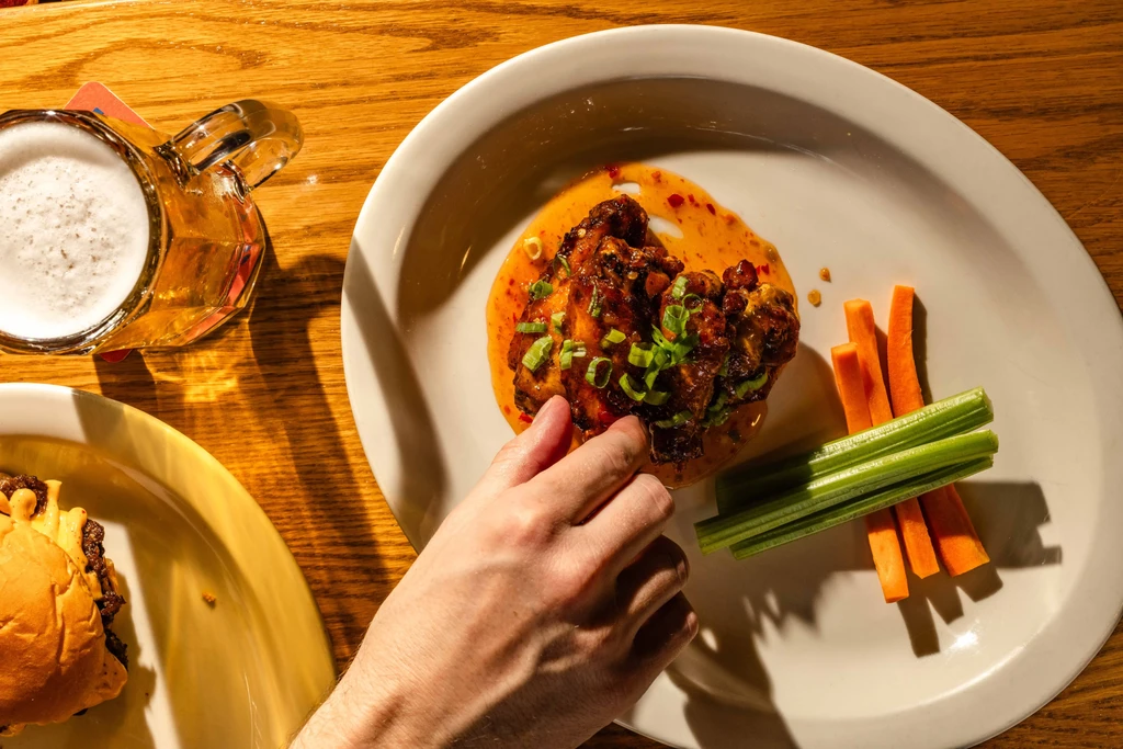 Hand reaches for sau sauced chicken wings, carrots, celery, burger, and beer.