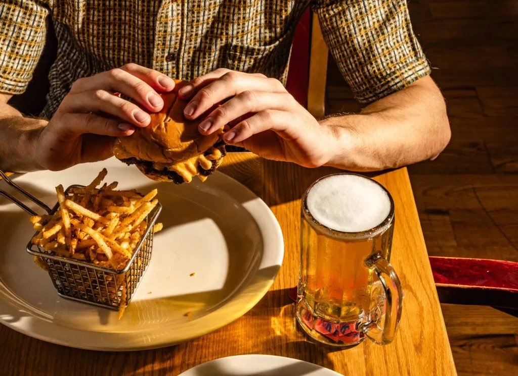 Hands hold a cheeseburger, seasoned fries, and frothy beer.