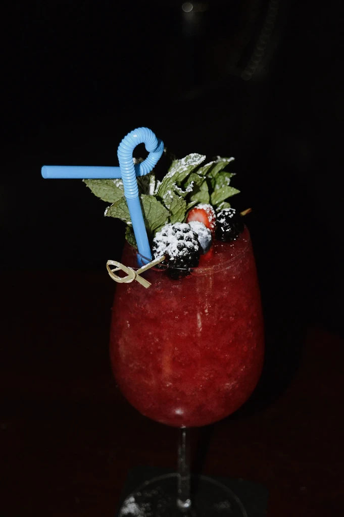 Slushy red drink in wine glass with mint, berries, blue straw.