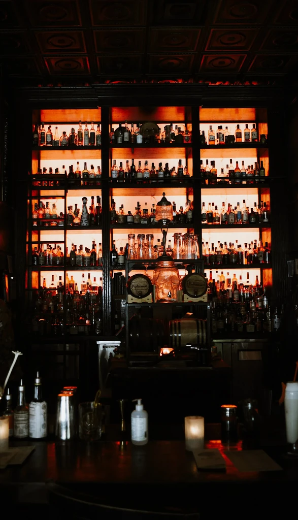Dark, well-stocked bar with orange backlit shelves, bottles, shakers, glasses.