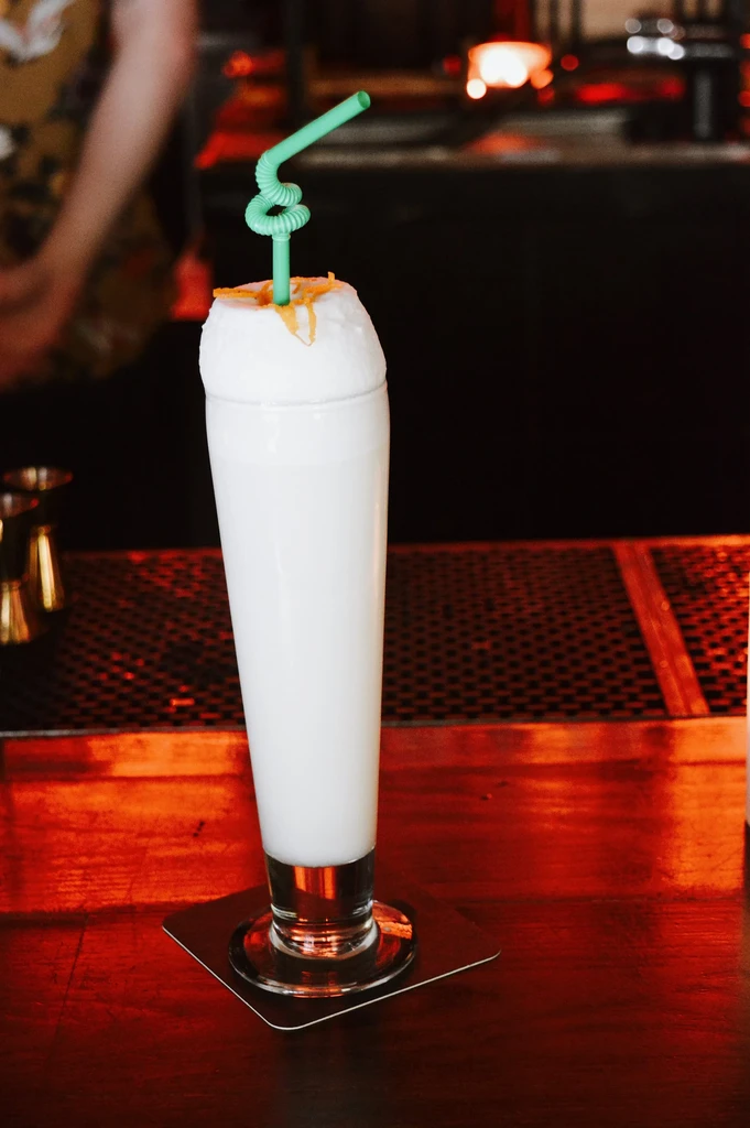 Frothy white cocktail with green straw and orange zest on a bar.