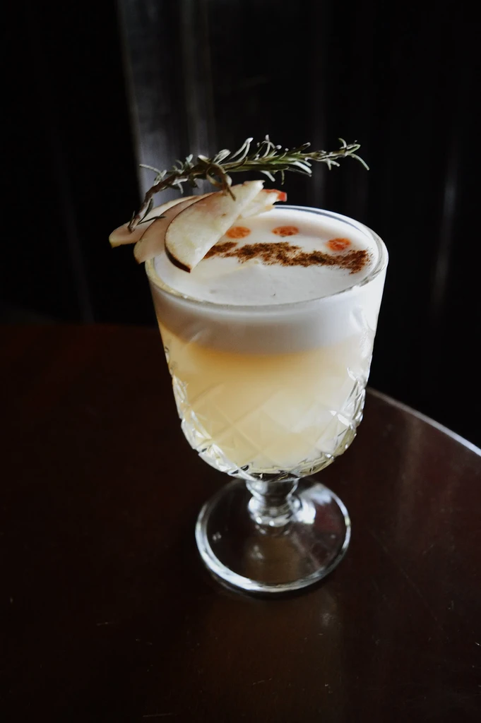Layered cocktail with foam, garnished with apple, rosemary, and spice.