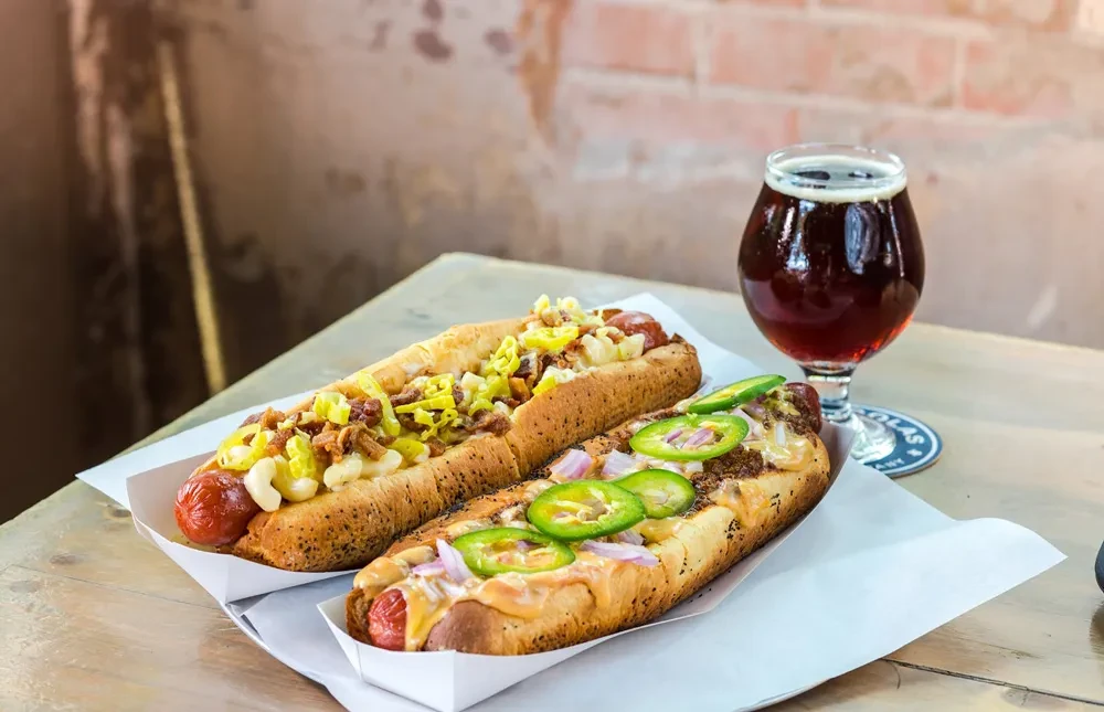 Gourmet hot dogs topped with chili, mac and cheese, next to a dark beer.