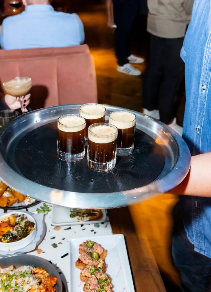 Server holds tray with four foamy dark drinks in a restaurant.