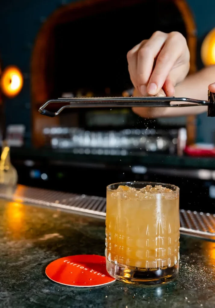 Hand grating spice over amber cocktail with CLIFTON CLUB coaster.