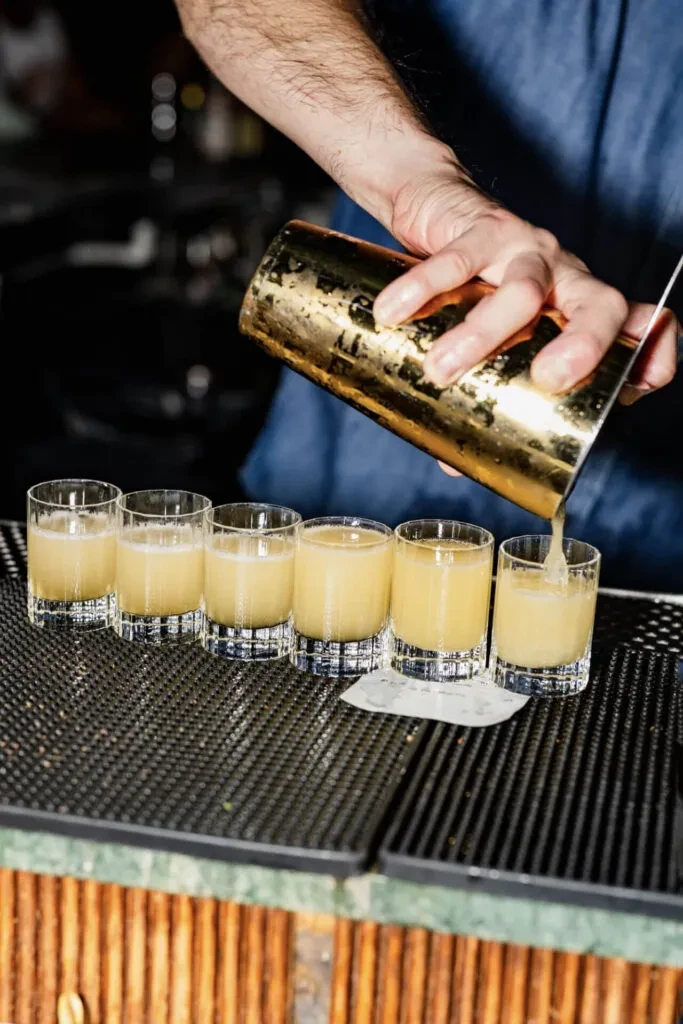 Bartender's hands pour yellow liquid from shaker into small glasses.