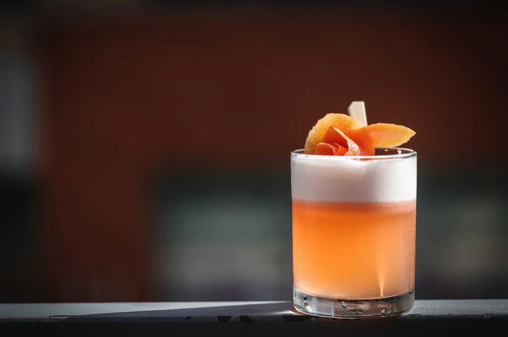 Orange-peach layered cocktail with frothy foam and citrus peel garnish.