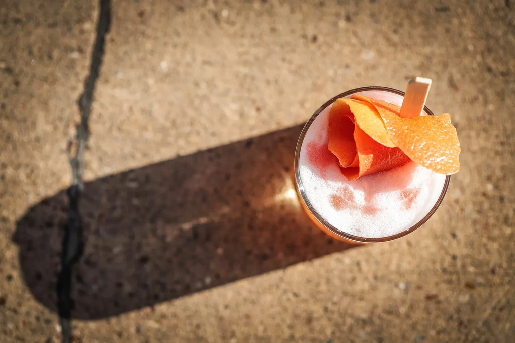 Overhead: pink-orange cocktail with foam, curled citrus peel, sunlight.