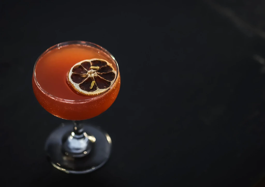 Reddish-orange cocktail in coupe glass, dried citrus garnish, dark background.