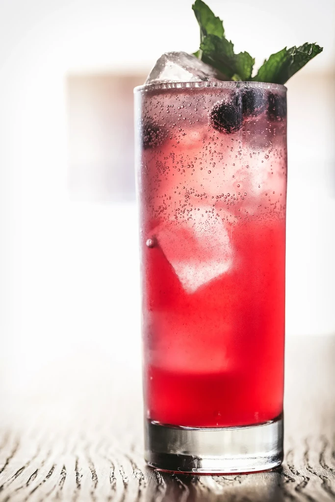 Tall glass of sparkling red drink with ice, blueberries, mint.