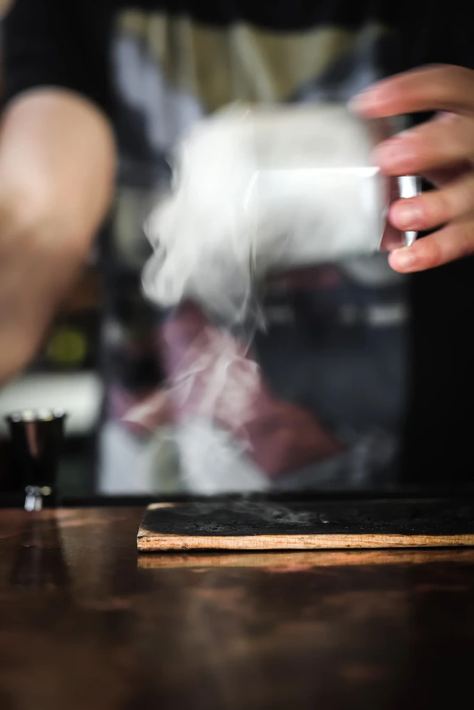 Hands hold a clear object with dramatic smoke rising over a bar.