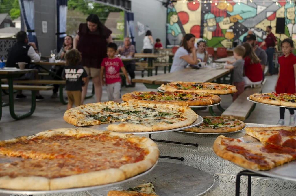Six diverse pizzas on tiered stands in a lively outdoor restaurant.