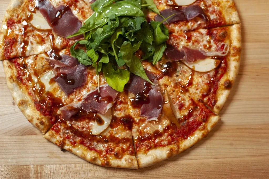 Overhead, sliced gourmet pizza with prosciutto, pear, arugula, balsamic.