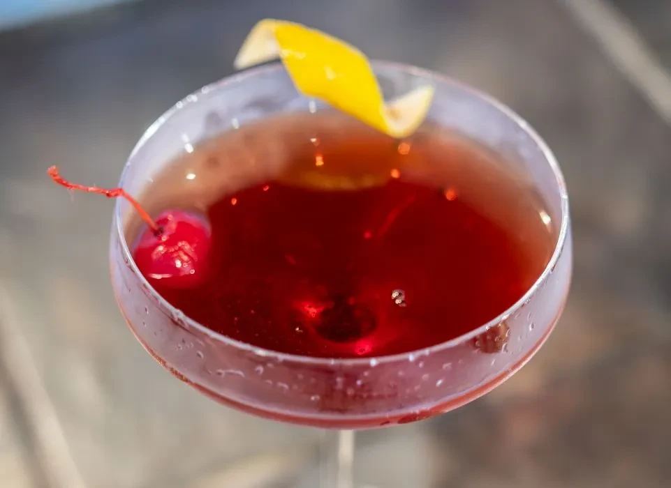 Ruby red cocktail with a cherry and lemon peel in a coupe glass.