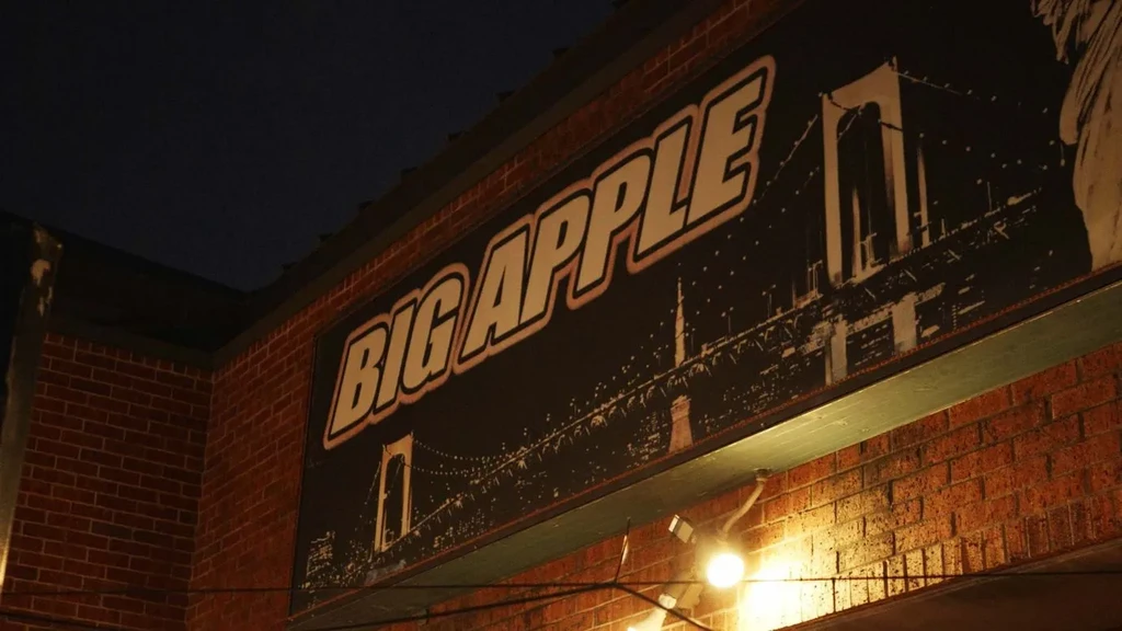 Night view: illuminated "BIG APPLE" sign and bridge on brick wall.